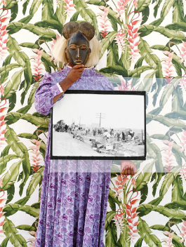 A color photograph of a Black woman in a full-length purple patterned dress, standing in front of floral wallpaper and holding a West African mask over her face and a large vintage black-and-white photograph.