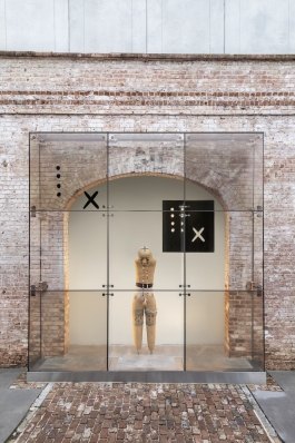 A mannequin without arms or feet sticks its head out forward, tilted downward. The figure is painted with designs and adorned with a necklace and is encased in a tall clear case made of see-through square panes. The  figure stands in a large brick archway, with a brick pathway leading to the clear case.  