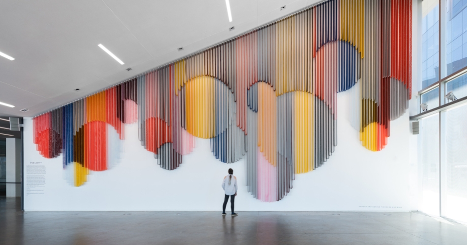 A visitor standing in a spacious lobby and looking up at a monumental hanging wall sculpture made from bands of colorful coated mesh fabric draped in various lengths to create series of interlocking circular forms. 