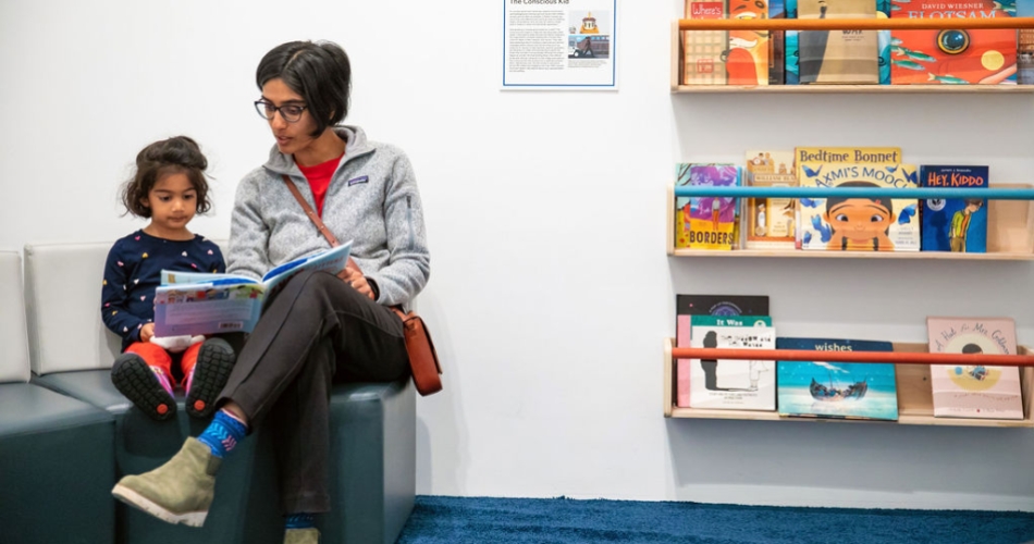A parent reads a picture book with their child with shelves of picture books next to them