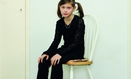 A color photograph of a light-skinned girl with brown pigtails wearing a black shirt and pants and red socks sitting on a chair, positioned at an angle and gazing at the viewer.