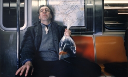 A color photograph of a middle-aged with pale skin, dark hair, and glasses holding a plastic bag containing a goldfish in water on the New York City subway.