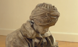 A bronze sculpture of a man in robes and a turban, looking at his hands, seated on the wooden floor of a gallery.