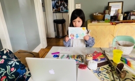 A young child holding up a crayon drawing to show on her laptop over a table full of colorful art supplies.