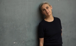 Anne Teresa De Keersmaeker, in a black top and blue jeans, leans against a gray wall.