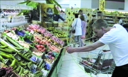 A video still shows the artist, a young white man in a white t-shirt, aiming a bow and arrow at a display of assorted vegetables in a grocery store.