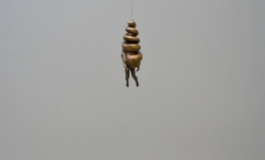 A small, bronze sculpture of a conical spiral with arms and legs suspended from a wire hovering over a slate disk.