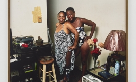 A color photograph shows two Black women posing together in cluttered living space. One raises her zebra-print dress to expose a prosthetic leg. The other is in a floral swimsuit. Both have their hair pulled back and smile toward the viewer.