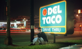 A color photograph of a young, light-skinned man sitting in front of a sign that reads "DEL TACO DRIVE THRU."