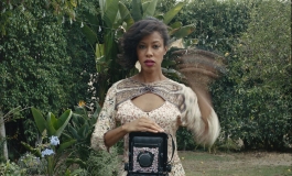 Woman with dark hair and a light colored flowery dress takes portrait with bedazzled 8x10 camera outdoors amongst blue flowers and green trees. Her left arm is blurred in motion.