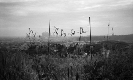 A grayscale photograph of the phrase "The Day-Sob-Dies" suspended between two thin poles in an overgrown field against the skyline of a city in the distance. 