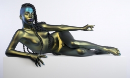 A life-size, hyper realistic bronze sculpture of a nude with long, thick hair in twists, lying on their side, propped up on one elbow with the other hand extended. The finish is a metallic green, except for blue fingernails and purple lipstick.