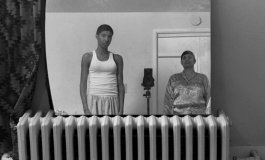 A black-and-white photograph shows the artist, a Black woman, and her mother in lounge wear and hair caps reflected in a mirror resting on a gas radiator.