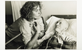 A black-and-white photograph of a light-skinned young woman leaning over the railing of a hospital bed, holding the hand of a frail and pale elderly woman who lays in the bed and looks up at her. 