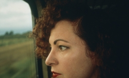 A color photograph shows the artist, a light-skinned woman with curly reddish hair and red lipstick, close- up in profile as she gazes out of a train window.