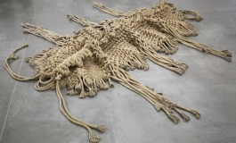 A sculpture made of very thick, beige rope or cordwoven together to resemble an abstracted inchworm on a concrete floor.