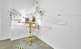 An elaborate mixed media installation occupying the corner of a white room and consisting of peeling white material, various yellow and orange materials, and a yellow metal tripod.