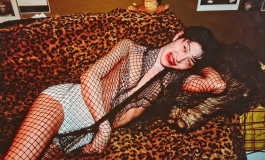 A color photograph of a Japanese, male-presenting young adult laying on their side on a leopard print sofa, smiling through smudged red lipstick, and wearing a black fishnet scarf over white underpants.