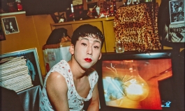 A color photograph of a Japanese adult with powdered makeup and red lipstick looking directly at the viewer in a cluttered room.