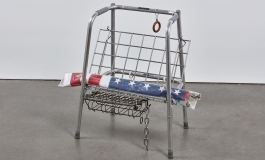 A sculpture made of an orthopedic walker and various salvaged metal works with a rolled up American flag wrapped in plastic laying across it.