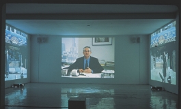 An installation of three videos projected onto three adjacent walls of a gallery. The central image shows a middle-aged white man in a suit seated in an office looking directly at the viewer, the others show a city storefront.