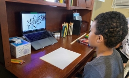 Young Black boy sat at a desk facing a piece of paper and laptop in front of him. 