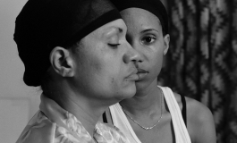 A black-and-white photograph shows the artist, a Black woman, looking directly at the viewer as she stands behind her mother, who is shown in profile and whose head obscures half of the artist's face. Both are wearing hair caps and a curtain is seen in the background.