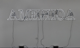 A white neon sculpture hung on a wall spells "AMERICA" in backwards capital letters.