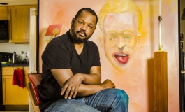 Artist Steve Locke sits in his studio in front of a large painting of a face with its tongue sticking out