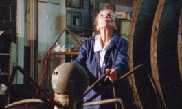 Inside an industrial studio, an elderly white woman looks up as she holds onto spider legs-like pipes from a metal sculpture.