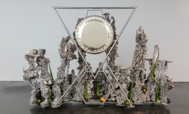 A silvery mixed-media sculpture with a hanging cymbal-like form surrounded by a number of standing, indistinct forms.