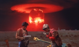 A color photograph depicts a scene of two Asian peasants, one played by the artist, posing with weapons in an open, devastated field as a red nuclear mushroom cloud blooms in the horizon.