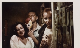 A grainy color photograph shows three light-skinned people in a hallway next to a wall of pinned-up images of nudes: a woman made up like a movie starlet and coyly posed, and two men in open collared shirts, one with a T-shirt underneath, holding theatrical masks and looking directly at the viewer.