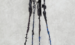 A sculpture comprised of a black wooden chair with elongated, spider-like legs that towers in height and is decorated with black feathers, black and silver tinsel, and hair.