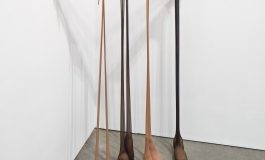 A sculptural installation consisting of four pairs of black and brown pantyhose suspended from a pole spanning the corner of a room. The feet of the pantyhose are filled with sand.