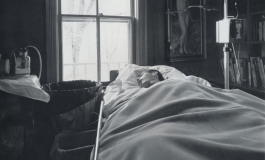 A black-and-white photograph of a man asleep under thick blankets in a home hospital bed, surrounded by an IV and other hospital equipment.