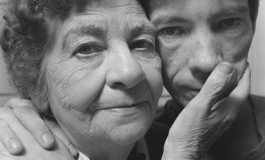 A black-and-white photograph of a light-skinned young man and his aging mother looking directly at the viewer, their faces touching.