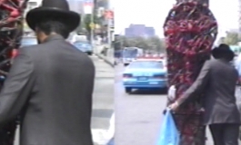Two side-by-side video stills show artist Nari Ward from behind as he pushes a large, towering sculpture on wheels down a crowded street wearing a suit and hat.