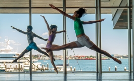 Dancers leap in front of large windows facing the Boston harbor