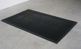 A sculpture of upright stainless-steel pins packed tightly onto a rectangular canvas on the floor, resembling a black rug.
