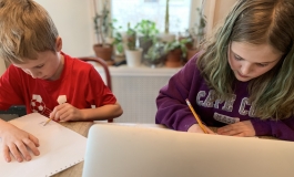 Two kids writing or drawing on a piece a paper on a table.