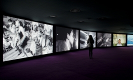 Six large video screens arranged side by side in a gentle curve fill a dark room. A woman stands before them, watching. They each display a different image, some black and white, including people dancing, some color. 