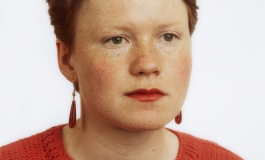 A color photograph portrait of a young woman with red hair, pale skin, red lipstick, and an orange sweater, shown in three-quarter profile.