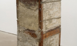 A sculpture consisting of a wooden chair almost completely within an unfinished block of cement and rebar.