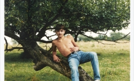A color photograph shows a light-skinned adolescent boy wearing blue jeans and no shirt seated on a low, leaning tree in an open field.