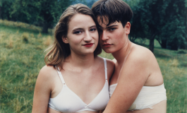 A color photograph of two light-skinned adolescents posed in an embrace in an open field, shirtless except for a bra and a binding bra and facing the viewer directly.