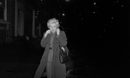 A black-and-white photograph of the artist on a very dark urban street, with blond hair and a dress skirt and blouse under a trench coat that she pulls closed around her neck.