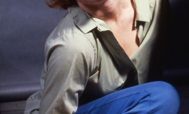 A color photograph of artist Cindy Sherman, a white woman, posed as Marilyn Monroe with blond hair and parted red lips, seated on the floor in jeans, boots, and a pale blouse.