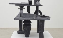 A sculpture of various ashy black, solid geometric stone and steel shapes stacked and balanced on top of one another.