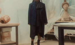 A medium-dark-skinned woman with braids and a dark dress, jacket, and boots stands in an art studio with several clay sculptures of heads. 